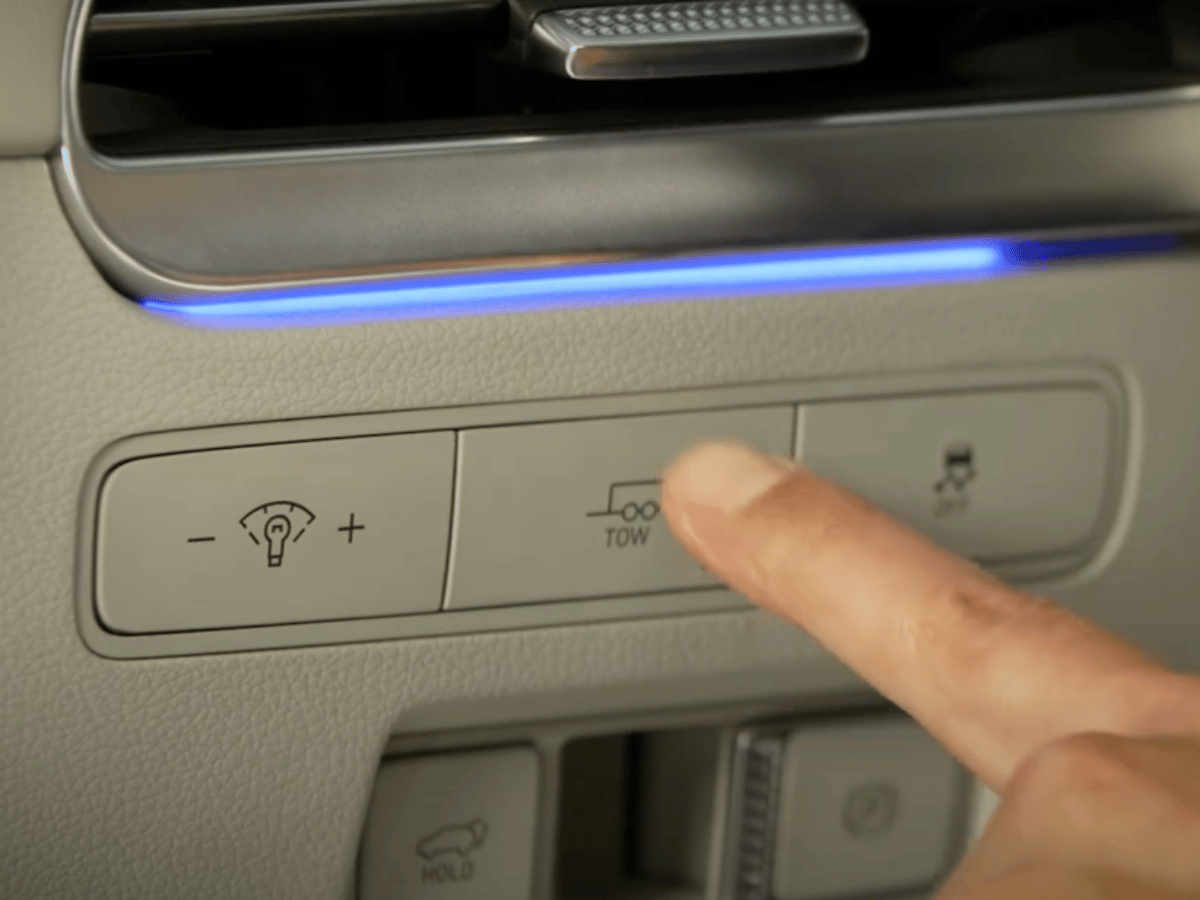 Driver's finger pressing the Tow Mode button in a 2025 Hyundai Palisade near headlight and traction control buttons