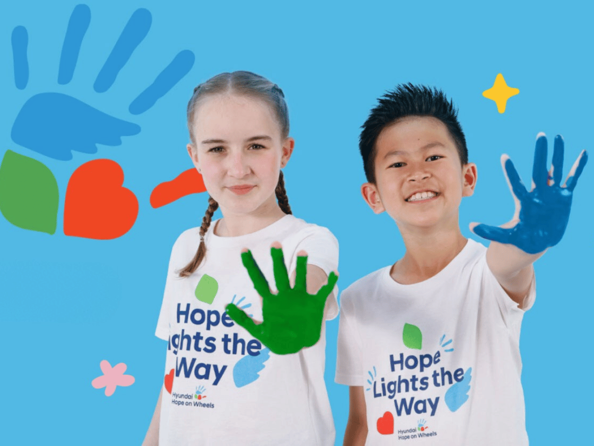 Two smiling children wearing “Hope Lights the Way” Hyundai Hope on Wheels t-shirts hold up painted hands — one with green paint, the other with blue. Colorful handprint and heart graphics appear in the background, symbolizing support and awareness for pediatric cancer research.
